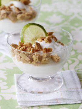 A glass cup filled with creamy yogurt, topped with crumbled cookies and garnished with a fresh lime slice, sits on a white napkin, with another similar dessert in the background.