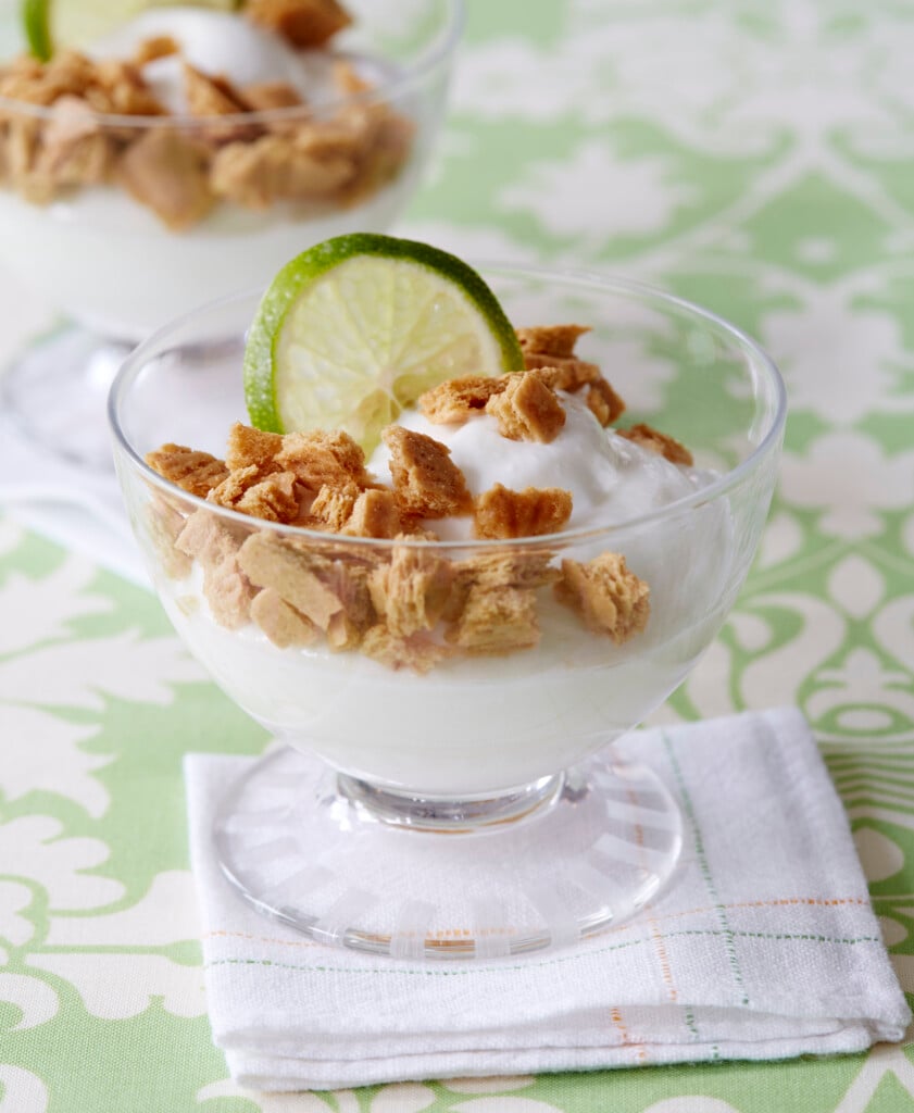 A glass cup filled with creamy yogurt, topped with crumbled cookies and garnished with a fresh lime slice, sits on a white napkin, with another similar dessert in the background.