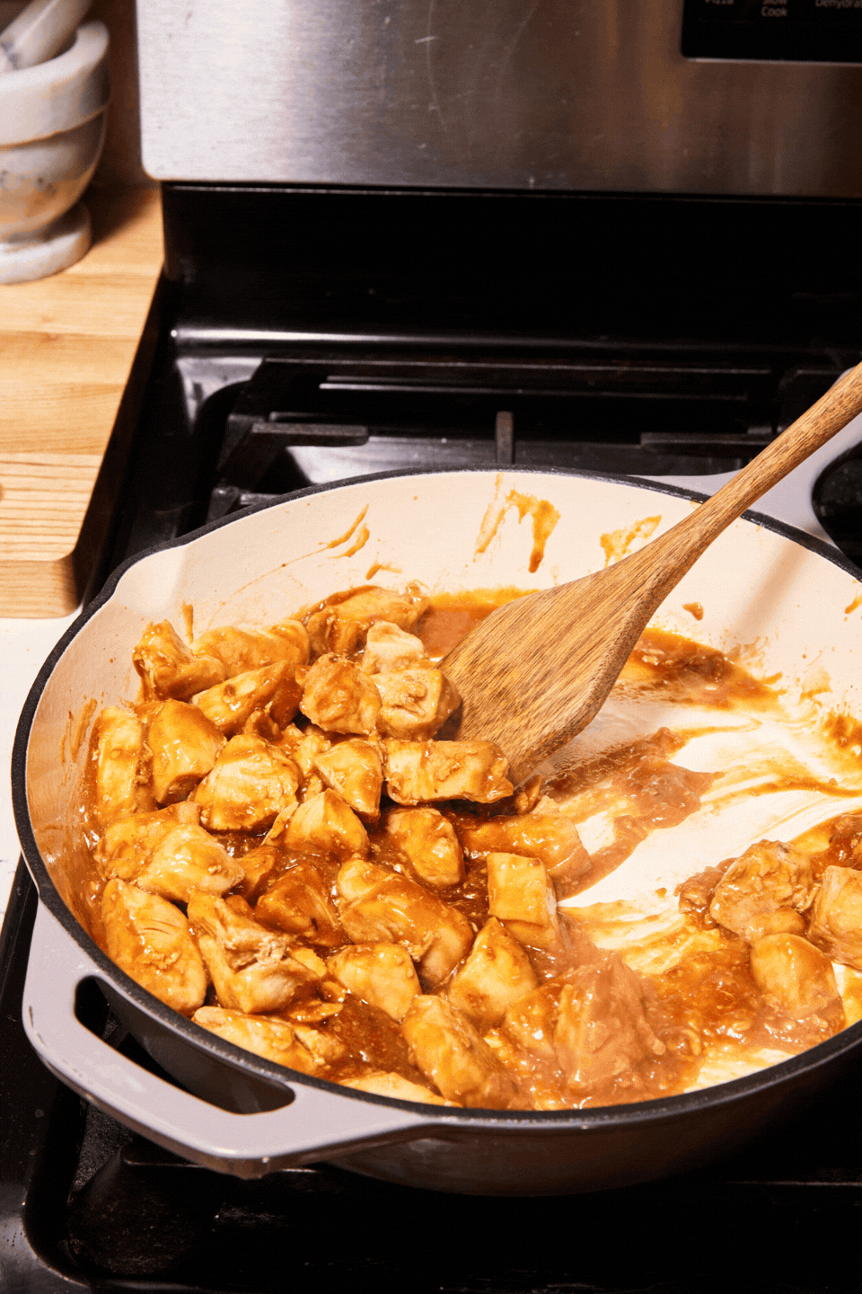 Chunks of General Joy’s Chicken cooking in a brown sauce in a white skillet on a stovetop, with a wooden spoon stirring the mixture. The stove and nearby wooden counter are partially visible.