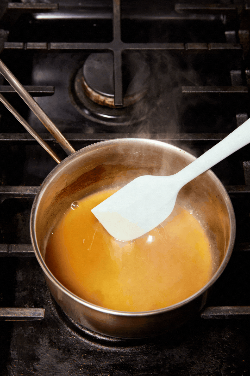A saucepan with a light brown liquid, reminiscent of General Joy’s Chicken sauce, is being stirred with a white spatula on a stovetop, with steam rising from the pot.