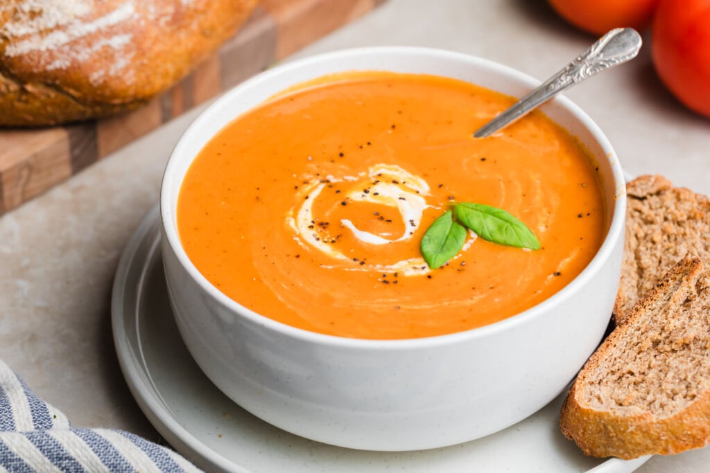 A bowl of Creamy Tomato Soup topped with a swirl of cream, black pepper, and fresh basil leaves, served alongside a slice of whole grain bread.