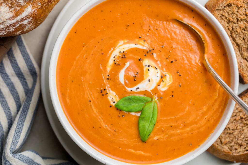 A bowl of Creamy Tomato Soup garnished with a swirl of cream, cracked black pepper, and two fresh basil leaves, with a spoon in the bowl and slices of bread on the side.