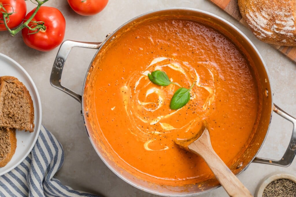 A pot of Creamy Tomato Soup garnished with basil leaves and a swirl of cream, with a wooden spoon resting in it. Slices of bread, fresh tomatoes, and a dish towel are arranged nearby for a cozy meal.