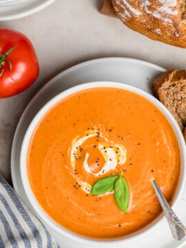 A bowl of Creamy Tomato Soup garnished with a swirl of cream, black pepper, and fresh basil leaves, served with slices of bread. A whole tomato and a striped napkin are nearby.