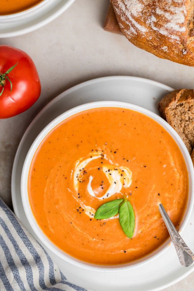 A bowl of Creamy Tomato Soup garnished with a swirl of cream, black pepper, and fresh basil leaves, served with slices of bread. A whole tomato and a striped napkin are nearby.