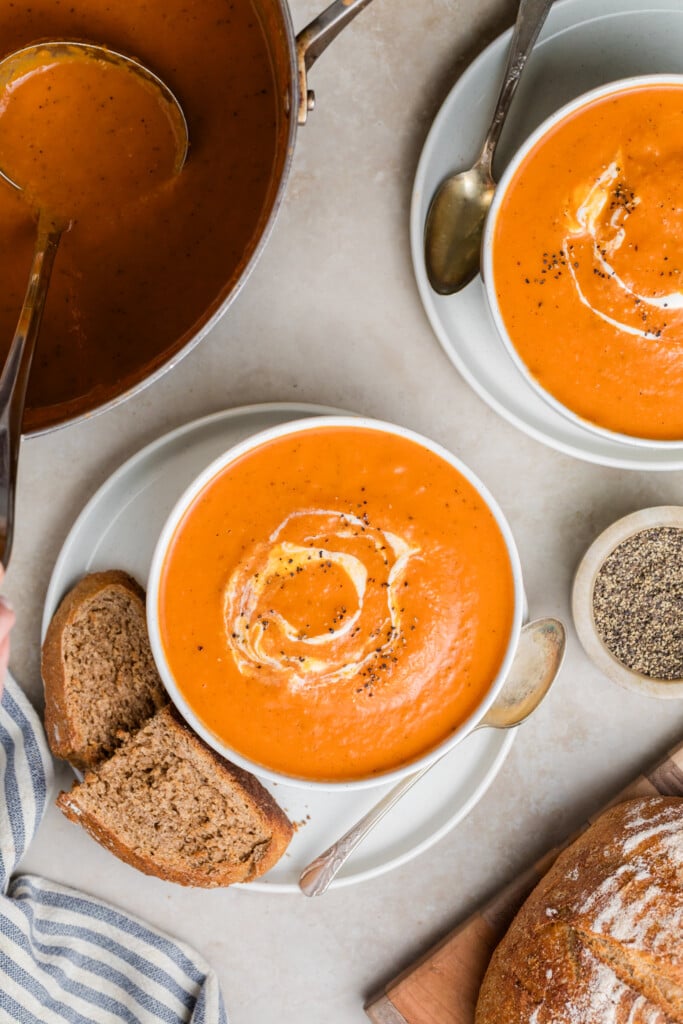 Two bowls of Creamy Tomato Soup with swirls of cream and cracked pepper, served alongside slices of brown bread. A pot of soup and a small bowl of ground pepper sit atop a light-colored table.