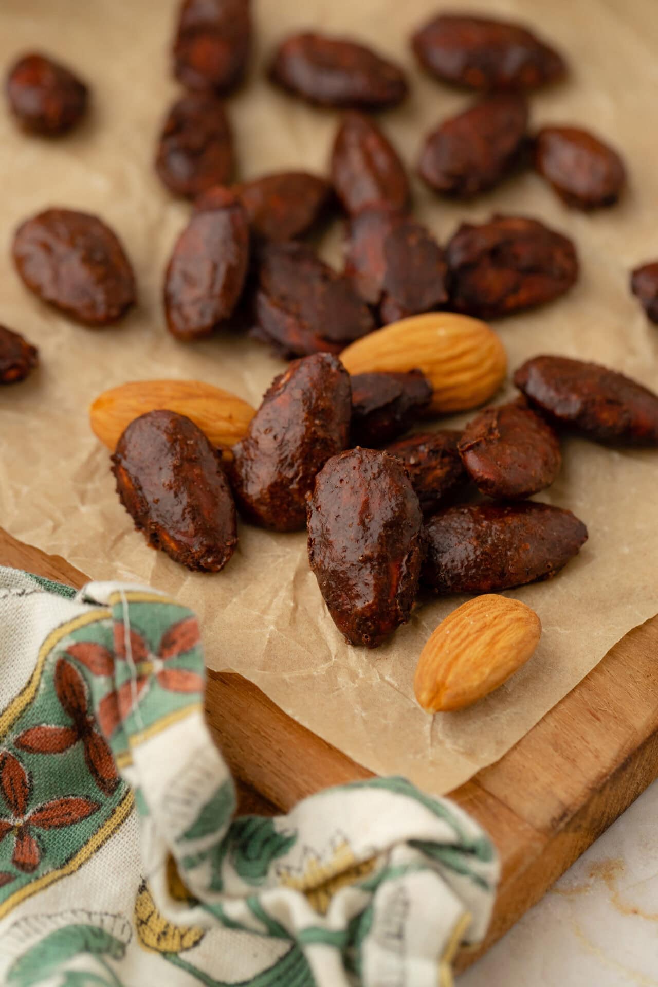 A close-up of dark chocolate almonds and plain almonds on parchment paper atop a wooden board, with part of a patterned cloth visible in the corner.