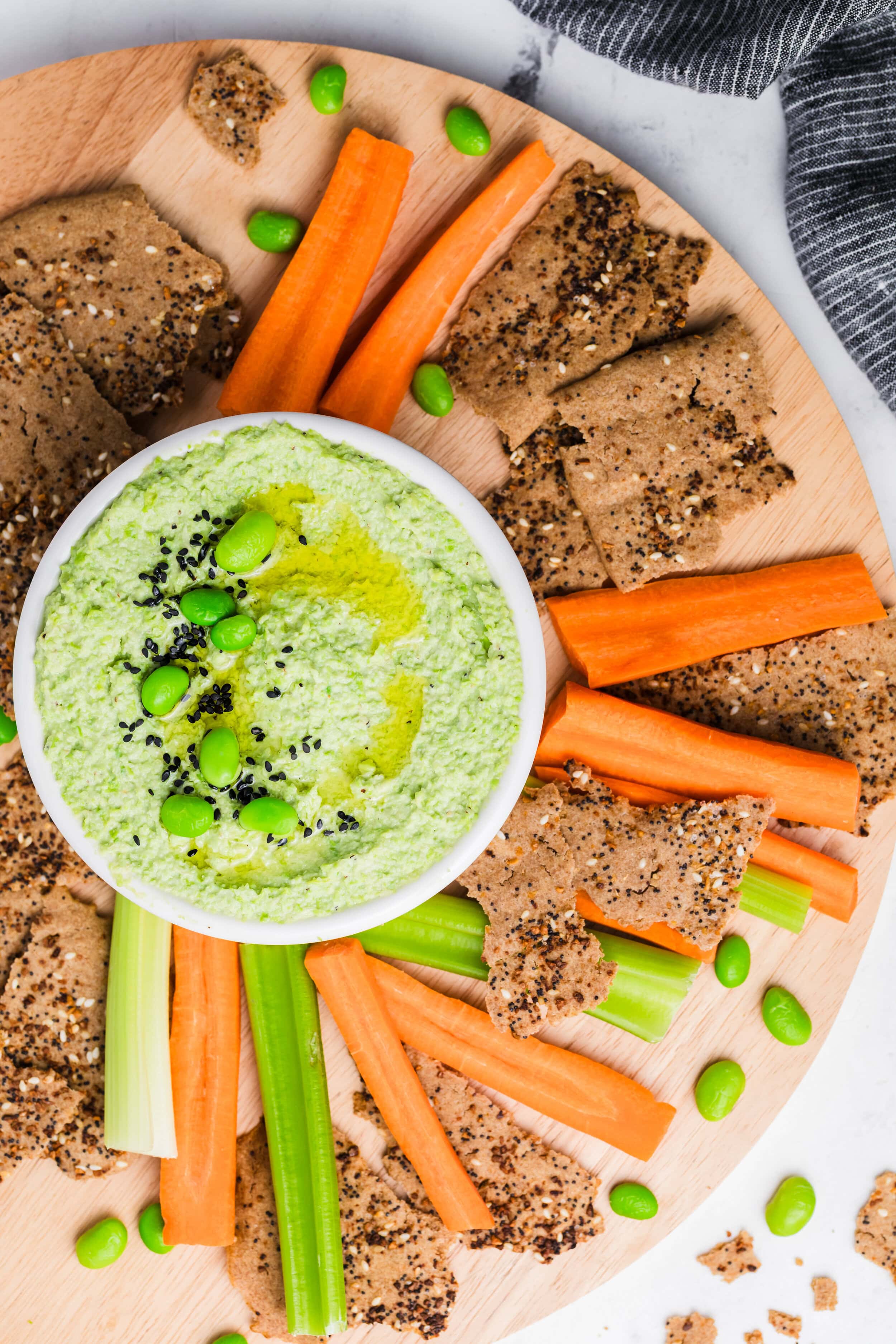 A round wooden platter features a bowl of creamy Edamame Hummus topped with edamame and black sesame seeds, surrounded by carrot and celery sticks, crackers, and scattered edamame beans.