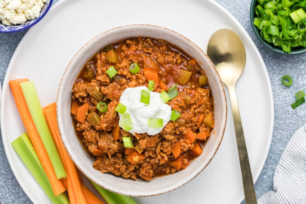 A bowl of Buffalo Chicken Chili topped with sour cream and chopped green onions, served on a white plate with celery and carrot sticks on the side. A gold spoon rests beside the bowl, with a small dish of green onions nearby.