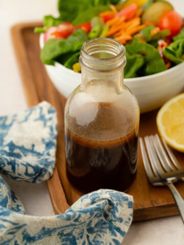 A glass bottle of balsamic vinaigrette sits on a wooden tray next to a fresh salad in a white bowl, a sliced lemon, a fork, and a blue patterned cloth napkin.