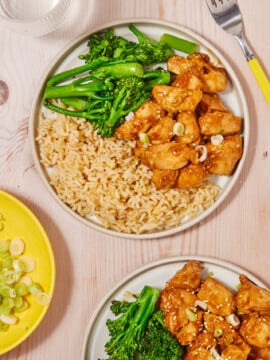 Two plates of food featuring General Joy’s Chicken—glazed chicken pieces topped with green onions—sit alongside servings of brown rice and steamed broccolini on a light wood table. A fork, glass of water, and a yellow plate with sliced green onions are also visible.