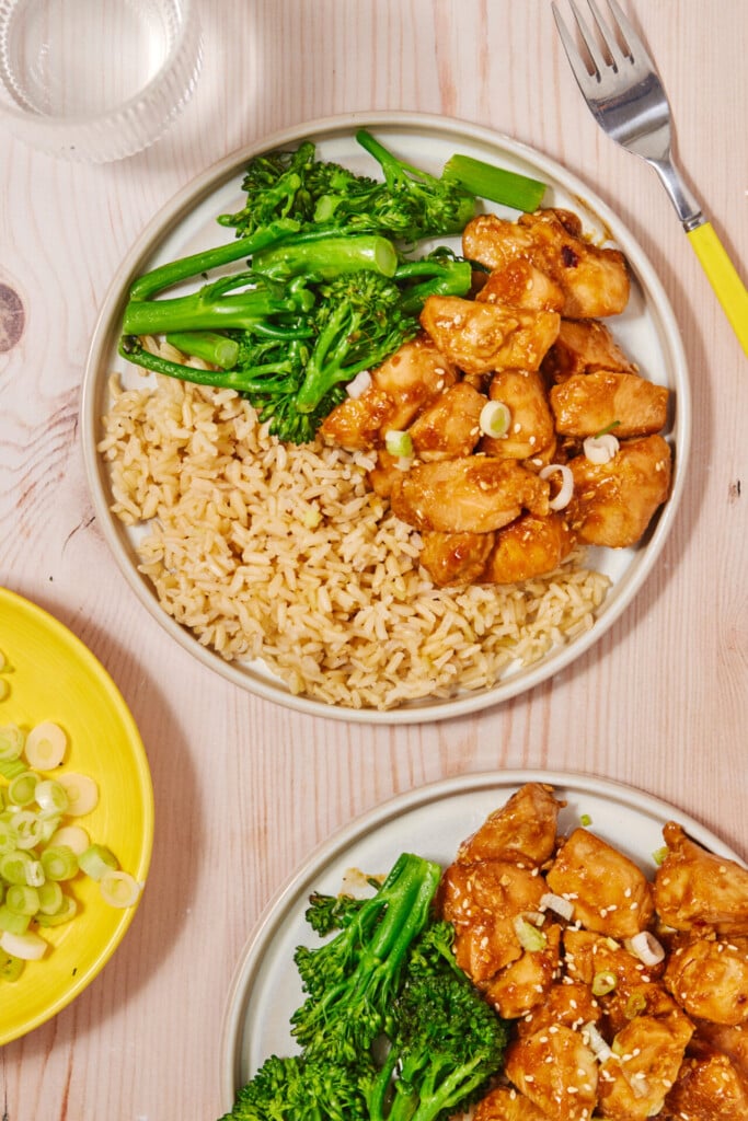 Two plates of food featuring General Joy’s Chicken—glazed chicken pieces topped with green onions—sit alongside servings of brown rice and steamed broccolini on a light wood table. A fork, glass of water, and a yellow plate with sliced green onions are also visible.