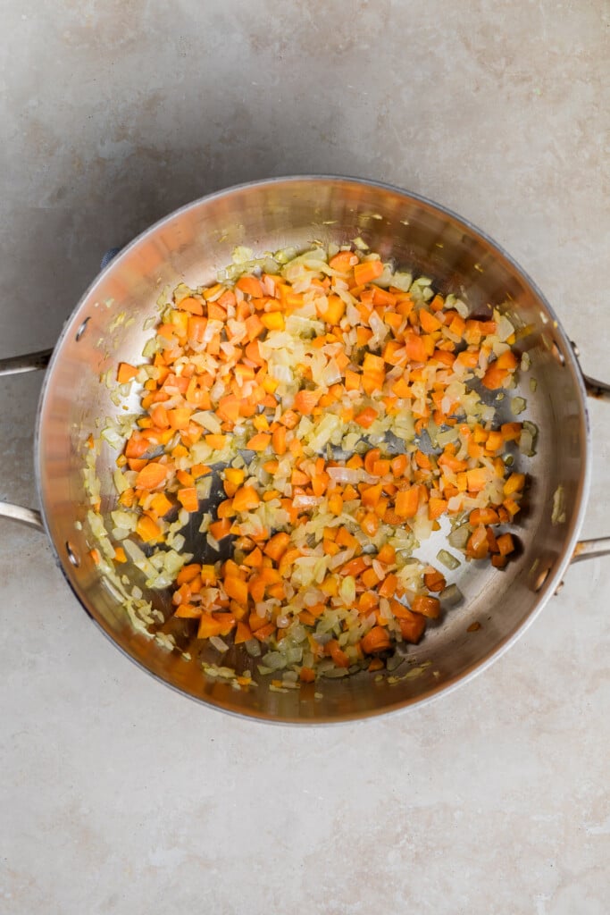 A stainless steel pan contains chopped carrots and onions being sautéed, with some pieces lightly browned—an essential base for a delicious Creamy Tomato Soup. The pan rests on a light-colored countertop.