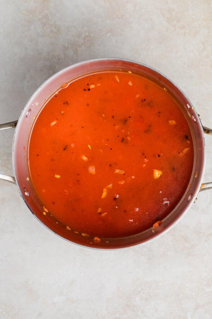 A stainless steel pot filled with creamy tomato soup, visible onion and seasoning pieces floating on the surface, placed on a light-colored countertop.