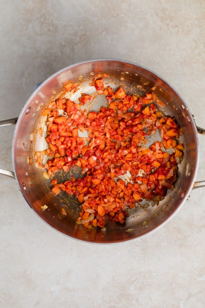 A stainless steel pot containing sautéed chopped onions and tomatoes, partially cooked for creamy tomato soup, sits on a light beige countertop.