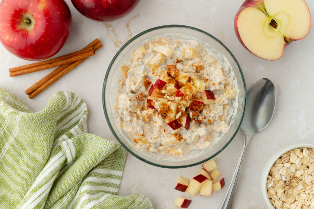 A bowl of Overnight Apple Pie Oats topped with apple chunks and cinnamon sits on a marble countertop, surrounded by fresh apples, cinnamon sticks, a green-striped towel, a spoon, and a small bowl of oats.