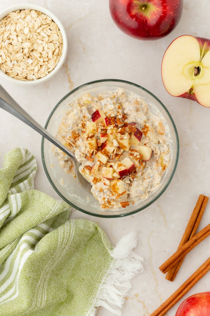 A bowl of Overnight Apple Pie Oats with chopped apples and cinnamon, surrounded by a green striped cloth, a whole apple, a halved apple, a bowl of oats, and cinnamon sticks on a light surface.