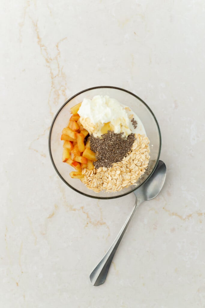 A glass bowl with Overnight Apple Pie Oats—yogurt, diced apples, chia seeds, oats, and milk—sits next to a spoon on a light marble surface.