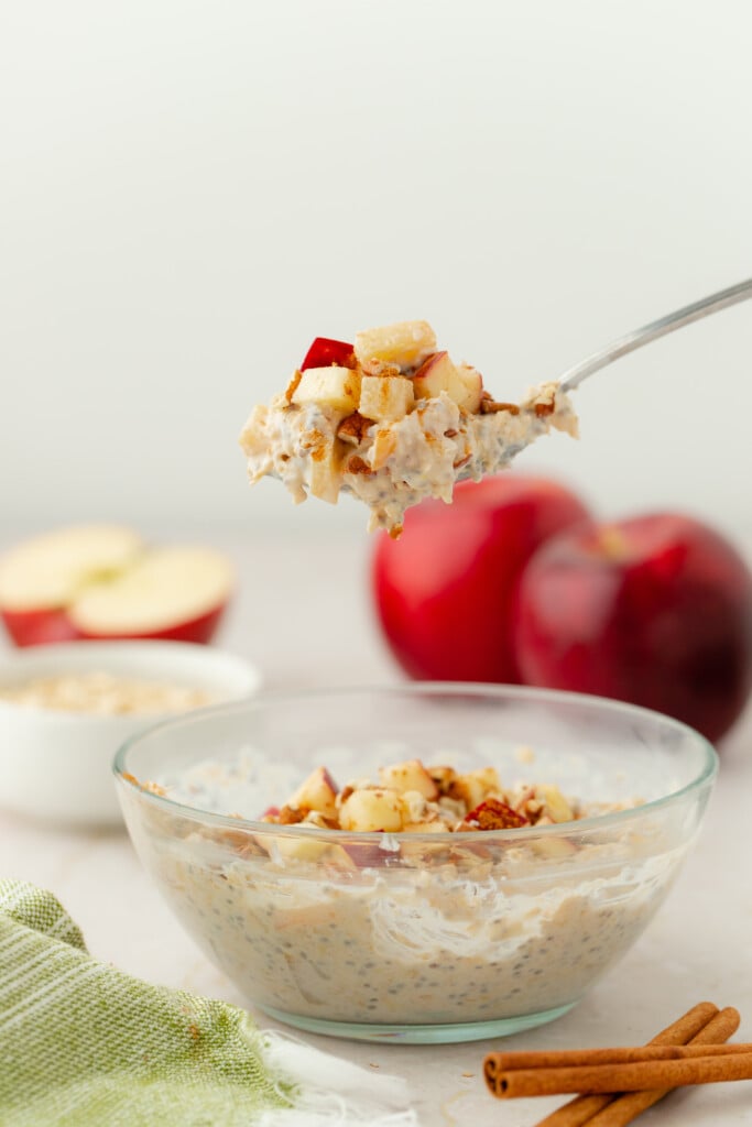 A spoonful of Overnight Apple Pie Oats with diced apples is held above a glass bowl filled with the same mixture. Two red apples, a green striped towel, and cinnamon sticks are visible in the background.