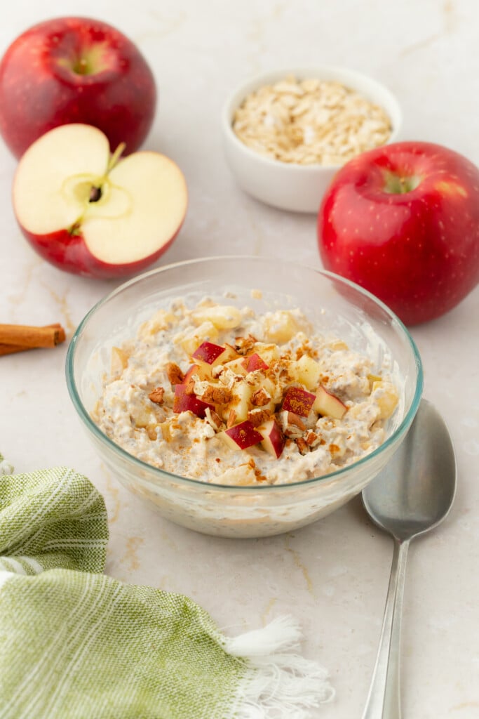 A glass bowl of Overnight Apple Pie Oats topped with chopped apples and nuts sits on a light surface, surrounded by whole and halved red apples, a bowl of oats, a spoon, a green cloth, and a cinnamon stick.