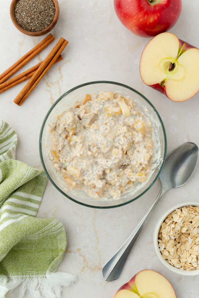 A bowl of Overnight Apple Pie Oats with apple pieces sits on a marble surface, surrounded by a spoon, apple halves, rolled oats, cinnamon sticks, ground pepper, and a green striped cloth.