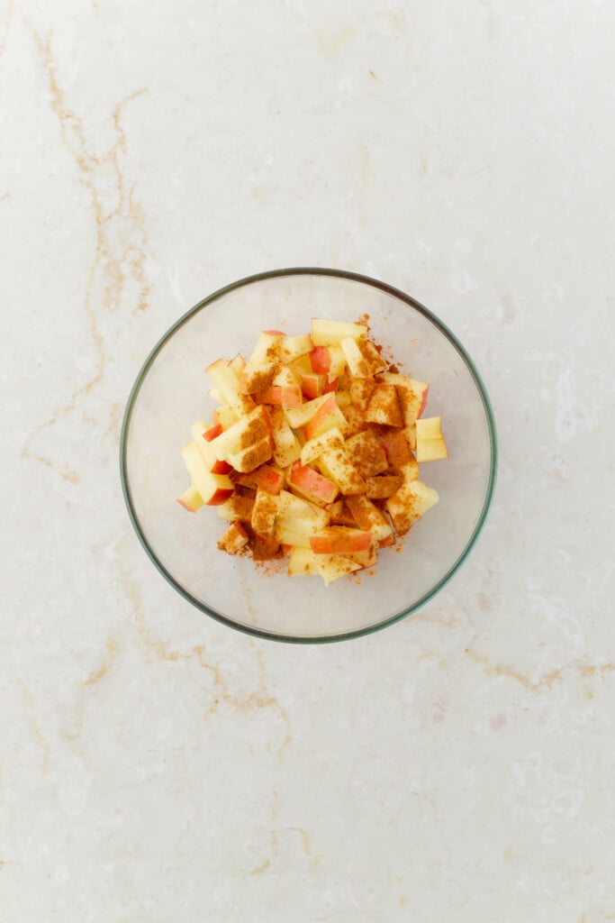 A glass bowl filled with chopped apples sprinkled with ground cinnamon sits on a light marble surface, perfect for mixing into Overnight Apple Pie Oats.