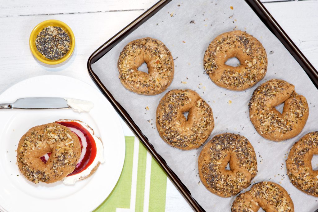 Protein Bagels on a baking sheet next to a protein bagel on a white plate.