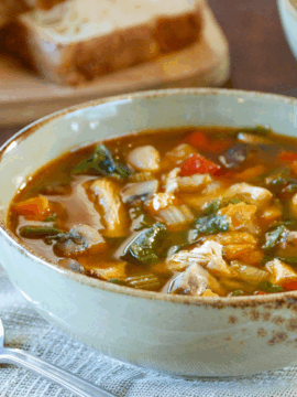 A bowl of Chicken & Vegetable Soup with visible greens, carrots, and pasta sits on a cloth napkin next to a spoon, with slices of bread on a wooden board in the background.