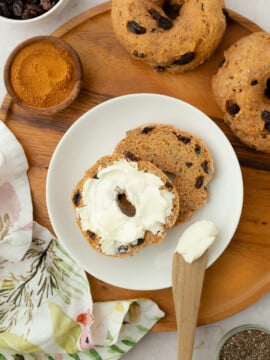 A sliced Cinnamon Raisin Protein Bagel on a white plate, one half spread with cream cheese. A wooden butter knife rests on the plate. Nearby are whole bagels, a bowl of cinnamon, raisins, and a patterned napkin.