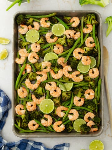 A One Sheet Mediterranean Shrimp with Charred Vegetables recipe featuring a baking sheet of cooked shrimp, green beans, broccoli, and lime slices. Extra veggies, lime wedges, and a blue checkered cloth rest nearby on the marble surface.