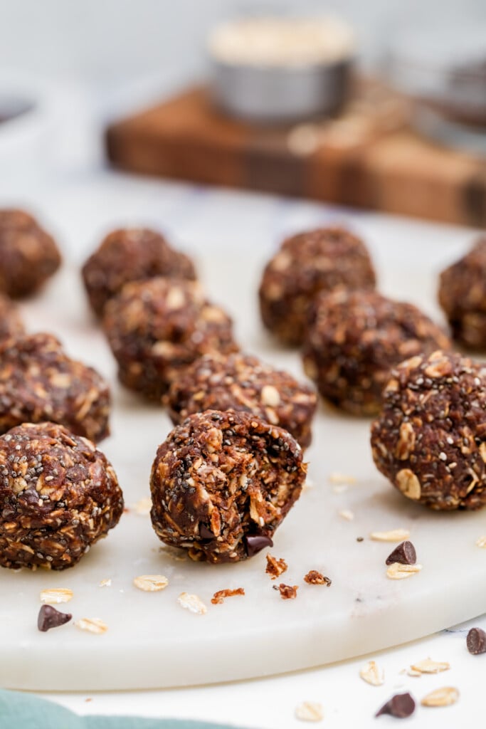 Several Chocolate Peanut Butter Energy Bites are arranged on a white marble surface, with one partially eaten in the front. Scattered oats and chocolate chips are visible around the bites.