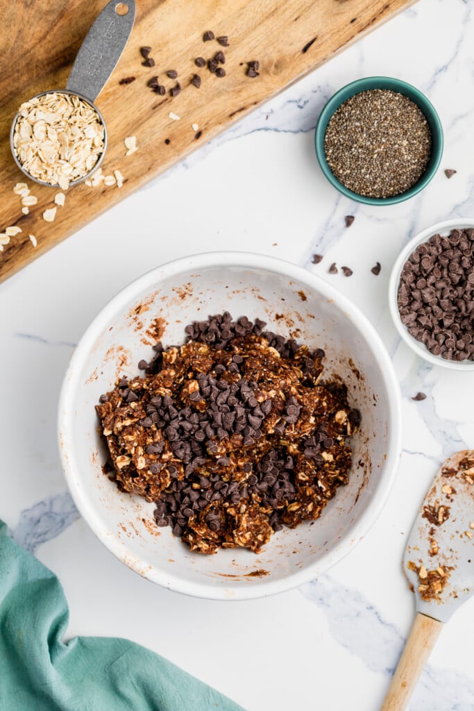 A white bowl filled with a mixture of oats, chocolate chips, and chia seeds sits on a marble counter—perfect ingredients for Chocolate Peanut Butter Energy Bites. Nearby are a wooden board with oats, chia seeds, and chocolate chips.