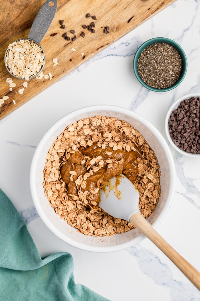 A white bowl filled with oats and peanut butter being mixed for Chocolate Peanut Butter Energy Bites. Surrounding the bowl are small bowls of chia seeds, chocolate chips, and a measuring cup of oats on a wooden board.