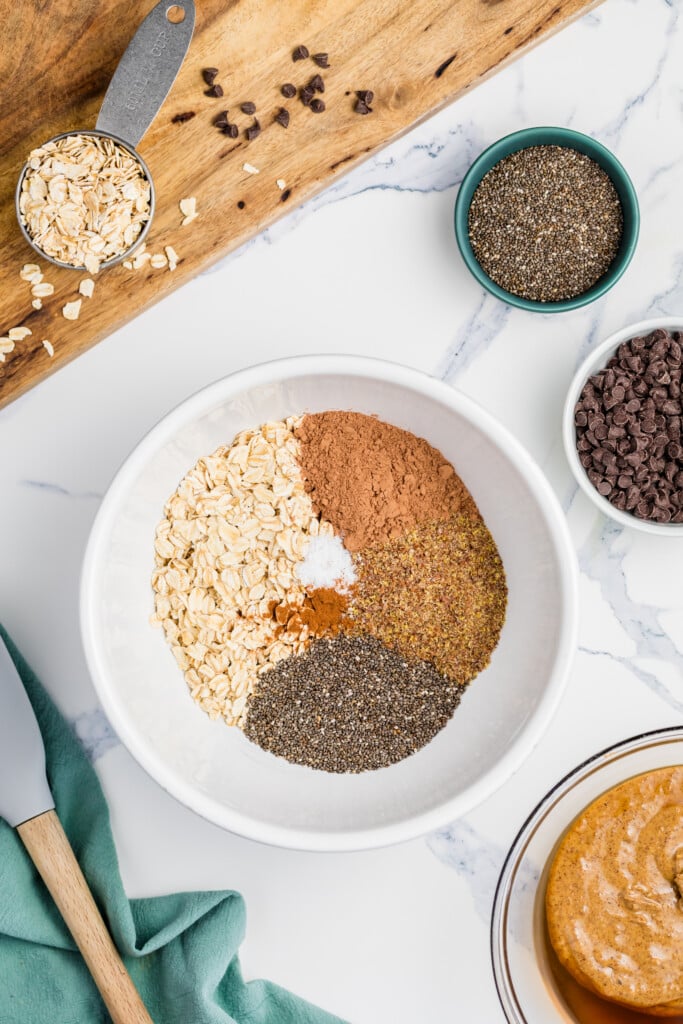 A white bowl with oats, cocoa powder, chia seeds, ground flaxseed, and salt arranged separately—perfect for making Chocolate Peanut Butter Energy Bites. Surrounding it are bowls with chocolate chips, nut butter, and a wooden spoon.