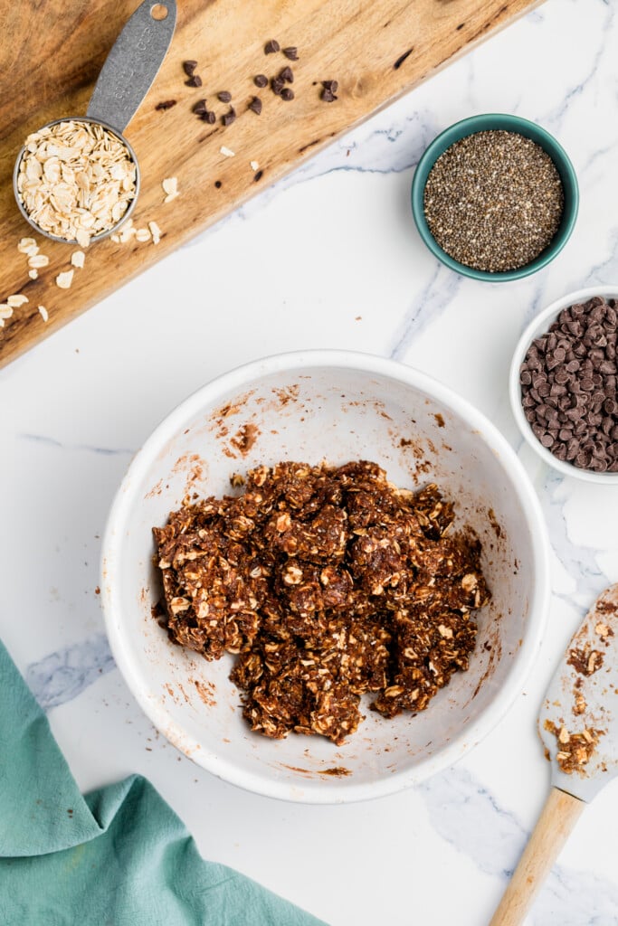 A white bowl with a mixture for Chocolate Peanut Butter Energy Bites—oats, chocolate chips, and chia seeds—sits on a marble countertop. Nearby are small bowls of chia seeds and chocolate chips, oats on a wooden board, and a spatula with some mixture.