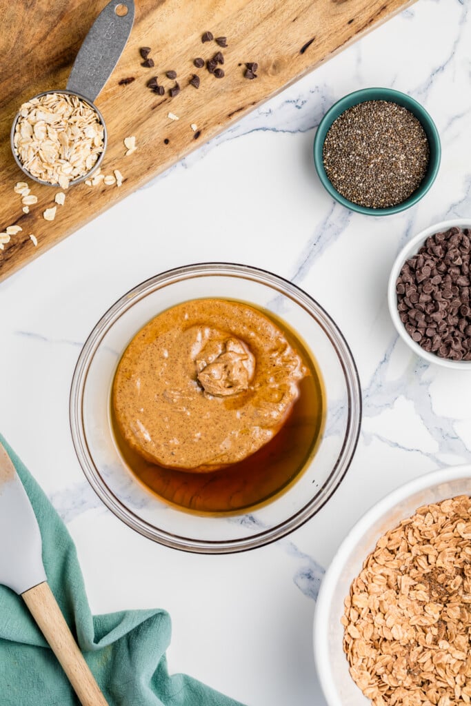 A glass bowl with almond butter and maple syrup on a marble counter, surrounded by bowls of oats, chia seeds, chocolate chips, and a measuring cup with oats—everything you need to make Chocolate Peanut Butter Energy Bites. A spatula and green towel are nearby.