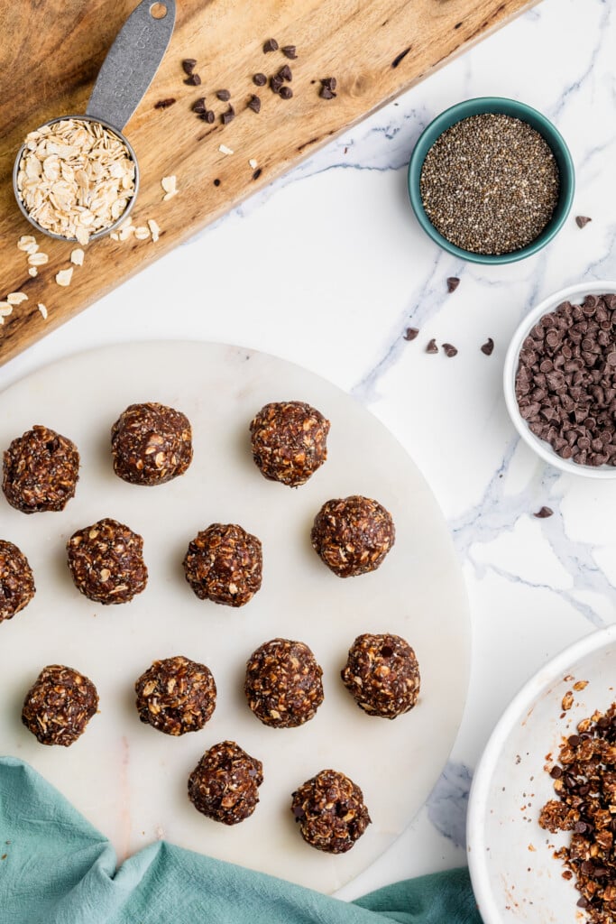 A marble surface displays Chocolate Peanut Butter Energy Bites arranged in rows, with bowls of oats, chia seeds, and chocolate chips, plus a wooden cutting board, all laid out for healthy snack preparation.