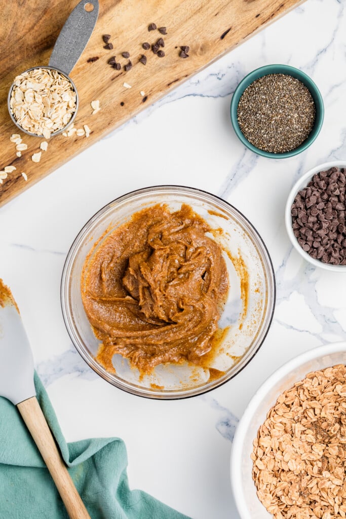 A glass bowl holds a thick brown batter for Chocolate Peanut Butter Energy Bites. Nearby are small bowls of chia seeds, oats, and chocolate chips, with a spatula and a green cloth on the marble surface.