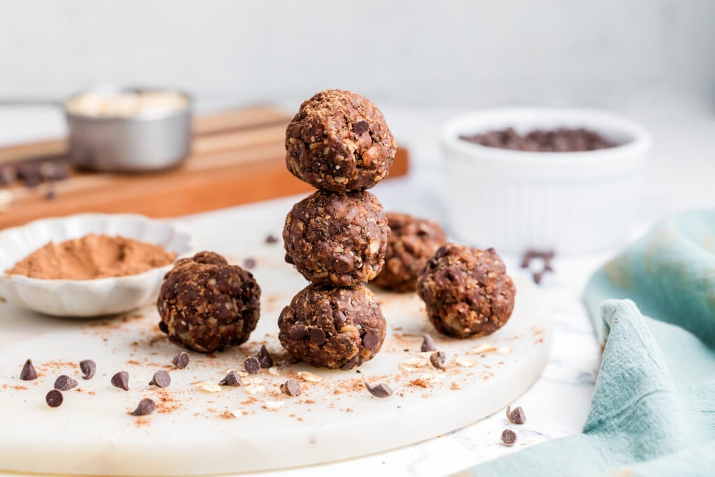 Four Chocolate Peanut Butter Energy Bites are stacked in a pyramid on a round marble board, with extra bites, chocolate chips, and cocoa powder scattered around. Blurred kitchen items are visible in the background.