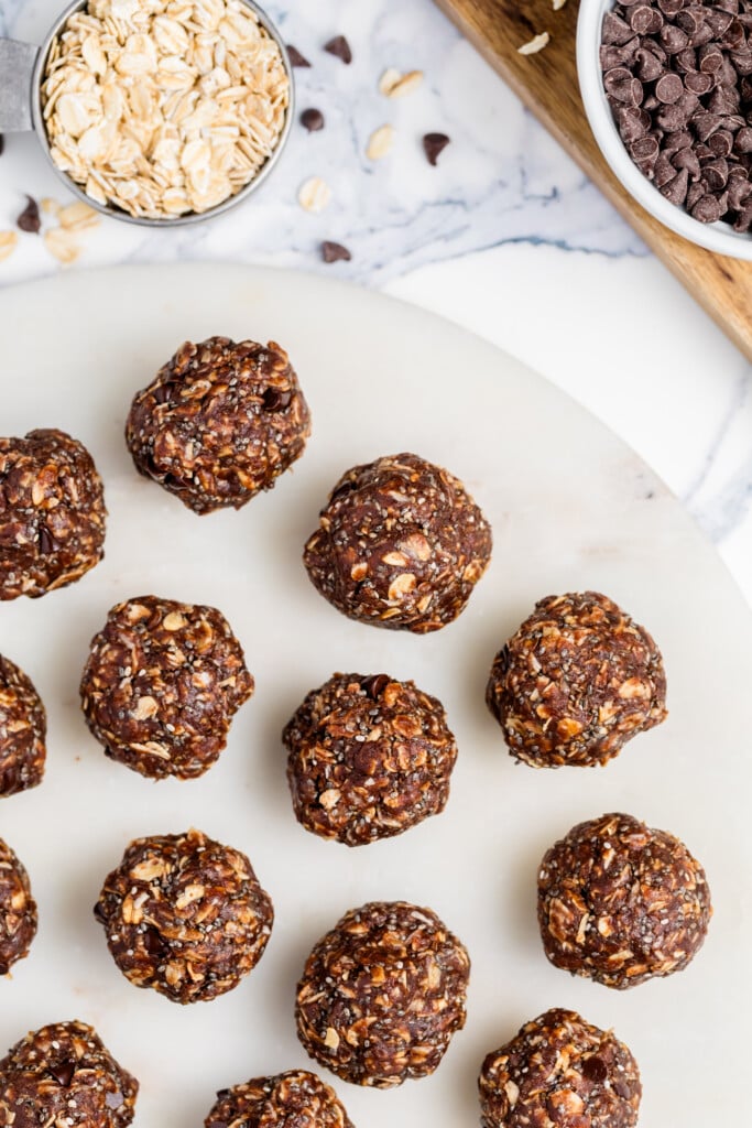 Round Chocolate Peanut Butter Energy Bites arranged on a white marble surface, with a bowl of oats and a bowl of chocolate chips visible in the background. Some loose oats and chips are scattered nearby.