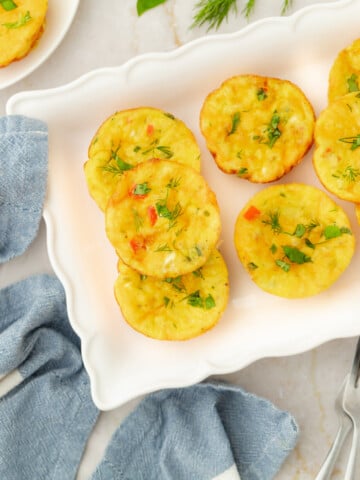 Mini egg frittata muffins with diced vegetables and herbs are arranged on a white plate. A blue cloth napkin and silver fork are nearby on the marble surface, with a few green herbs sprinkled around.