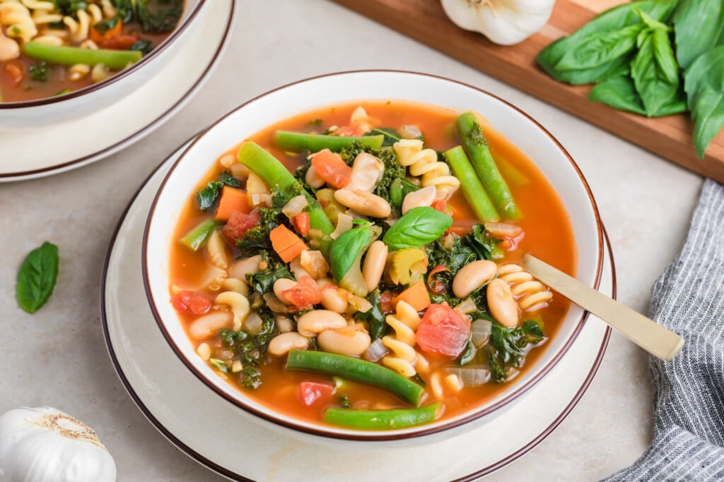A bowl of Superfood Minestrone with pasta, white beans, green beans, tomatoes, carrots, and leafy greens, garnished with fresh basil leaves. A spoon rests in the bowl, with garlic and basil nearby.