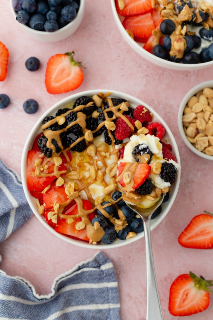 A PB and Berry Breakfast Bowl: creamy yogurt topped with strawberries, blueberries, blackberries, raspberries, chopped nuts, and a drizzle of peanut butter, served on a pink surface with fresh berries and a small bowl of nuts nearby.