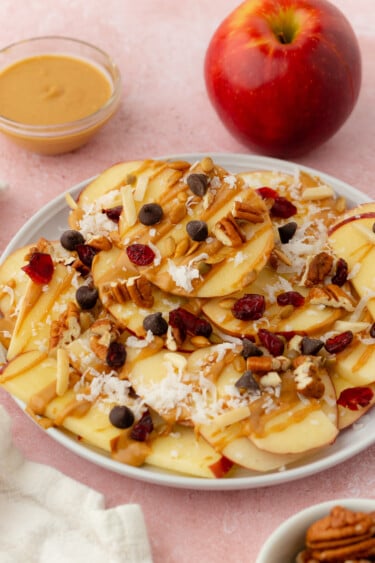 Single-Serve Apple Nachos feature sliced apples topped with shredded coconut, chocolate chips, nuts, dried cranberries, and a drizzle of peanut butter; a whole apple and a bowl of peanut butter sit in the background.