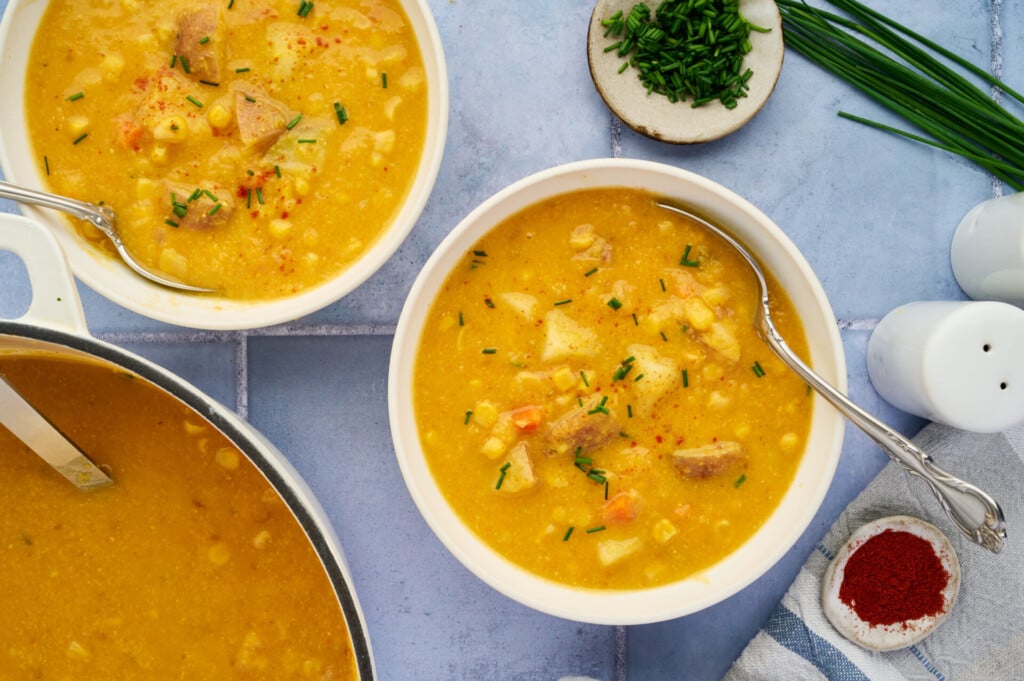 A bowl of thick yellow Corn Chowder with chunks of meat, corn, and veggies, garnished with chopped chives. Nearby are a pot of soup, a plate of chives, paprika, salt, and a striped towel with a spoon.
