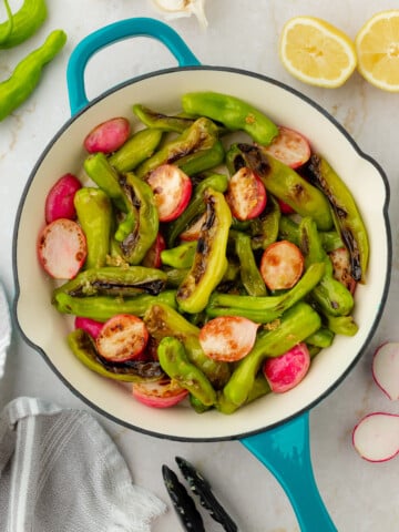 A close-up overhead shot of blistered shishito peppers and radishes in a teal skillet.