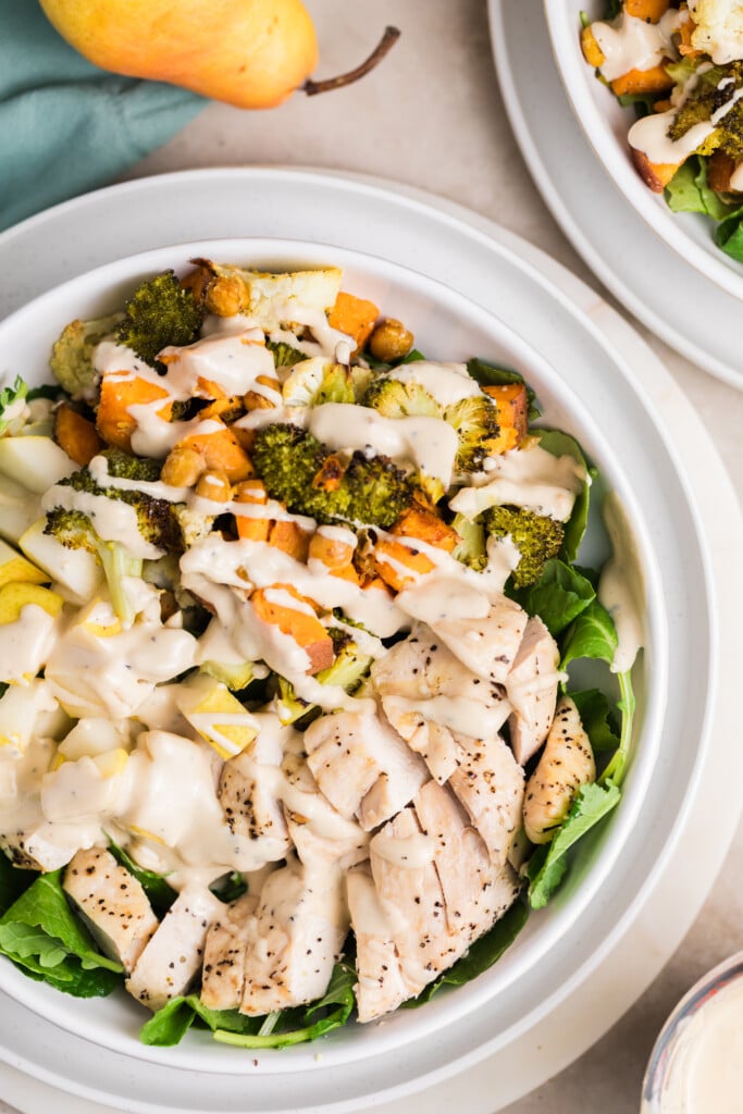 A Reboot Bowl featuring grilled chicken slices, roasted vegetables, leafy greens, and creamy dressing drizzled on top. Another bowl of salad and a pear are partially visible nearby.