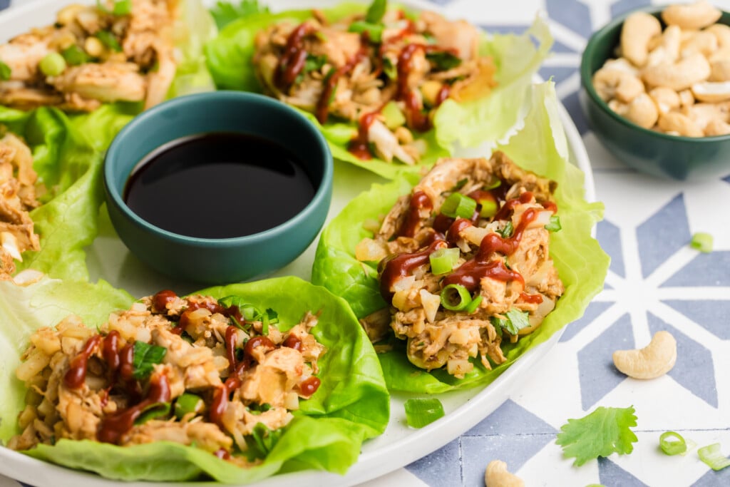 A plate of Rotisserie Chicken Lettuce Wraps filled with shredded chicken, chopped green onions, and sauce, served with a small bowl of soy sauce and a side of cashews. The dish is garnished with fresh cilantro.