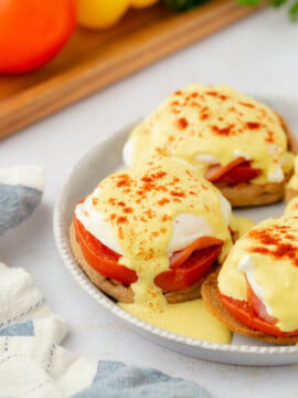 A plate of Eggs Benedict with poached eggs, sliced tomatoes, and English muffins, topped with creamy yellow pepper hollandaise sauce and sprinkled with paprika. A blue and white napkin rests beside the plate.