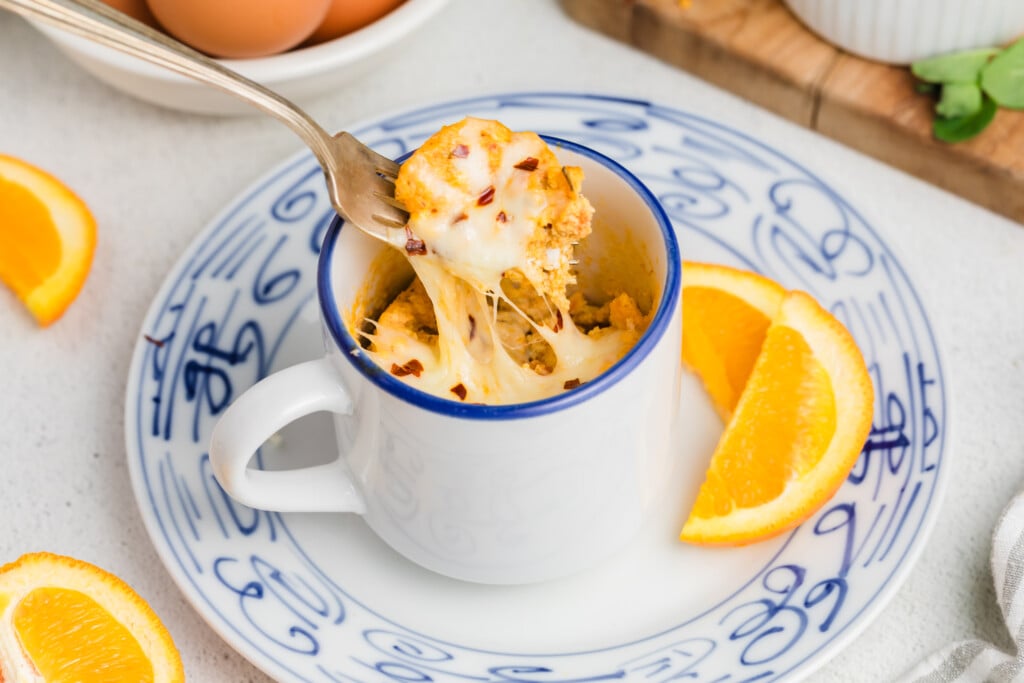 A fork lifts cheesy, baked egg from a white mug on a blue-patterned plate, with orange slices beside the mug. This Pizza Mug Omelet is surrounded by eggs and more orange slices in the background.